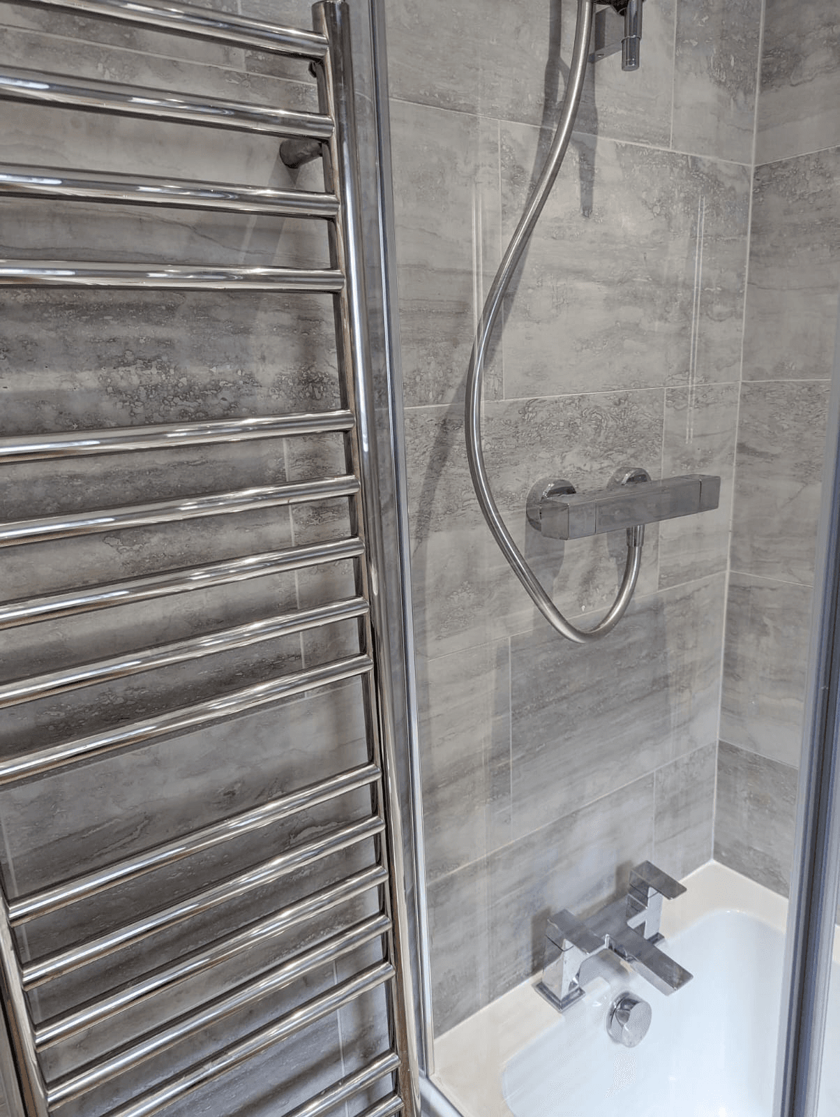 Polished chrome ladder heated towel rail next to bath and thermostatic bar shower