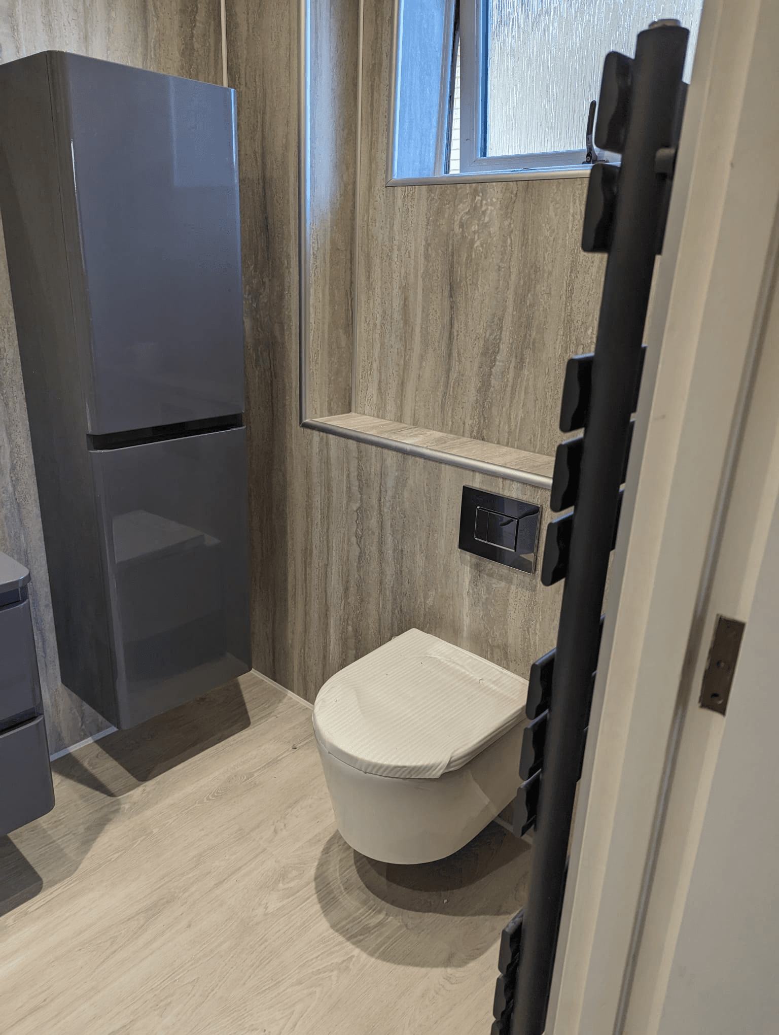 Grey tall storage unit beside wall-hung toilet with built-in shelf and wood-look floor tiles
