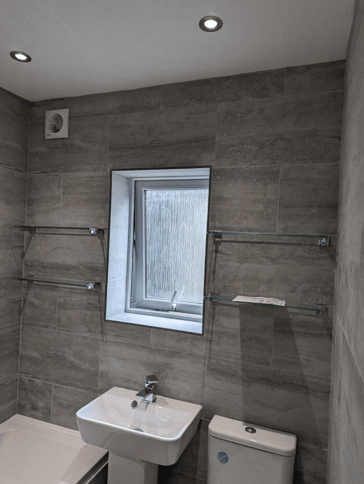 Grey tiled bathroom with bath, pedestal sink, toilet and chrome shelf rails beside frosted window