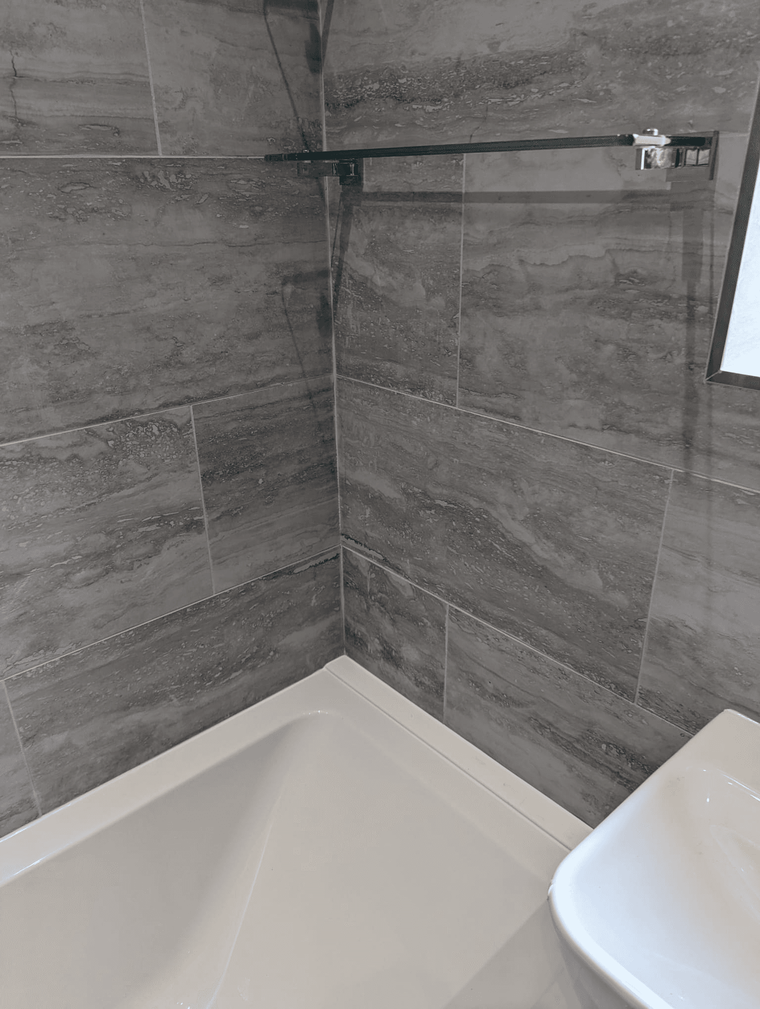 Bath corner surround with large-format grey marble-look tiles and glass bath shelf